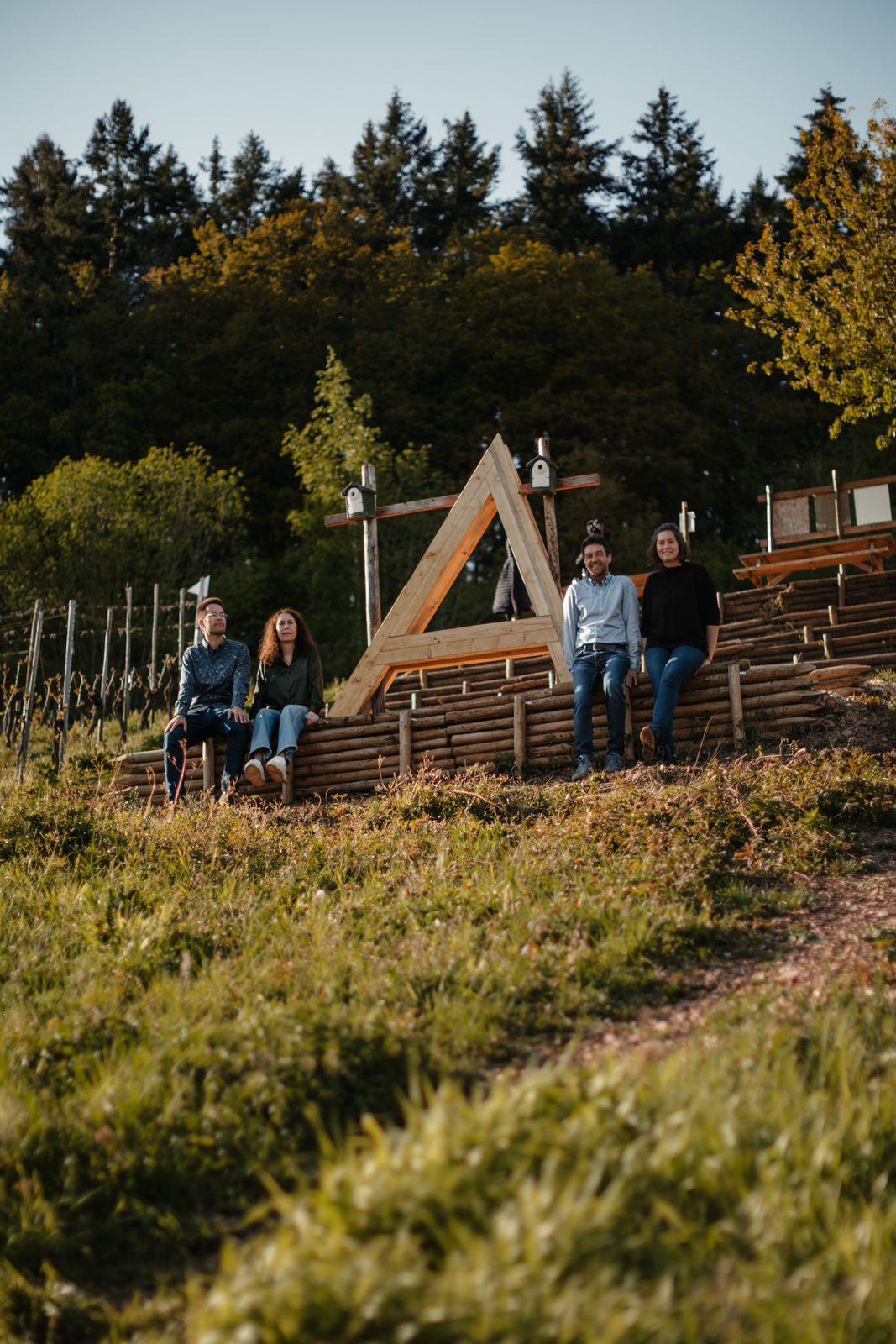 Vier Personen (Winzerfamilie Gindorf) sitzen entspannt an der Lagenliebe-Weinbergbühne oberhalb der Mosel; im Vordergrund Wiese, im Hintergrund Holztribüne und Wald – sonniger Tag im Weingut Günter Gindorf.