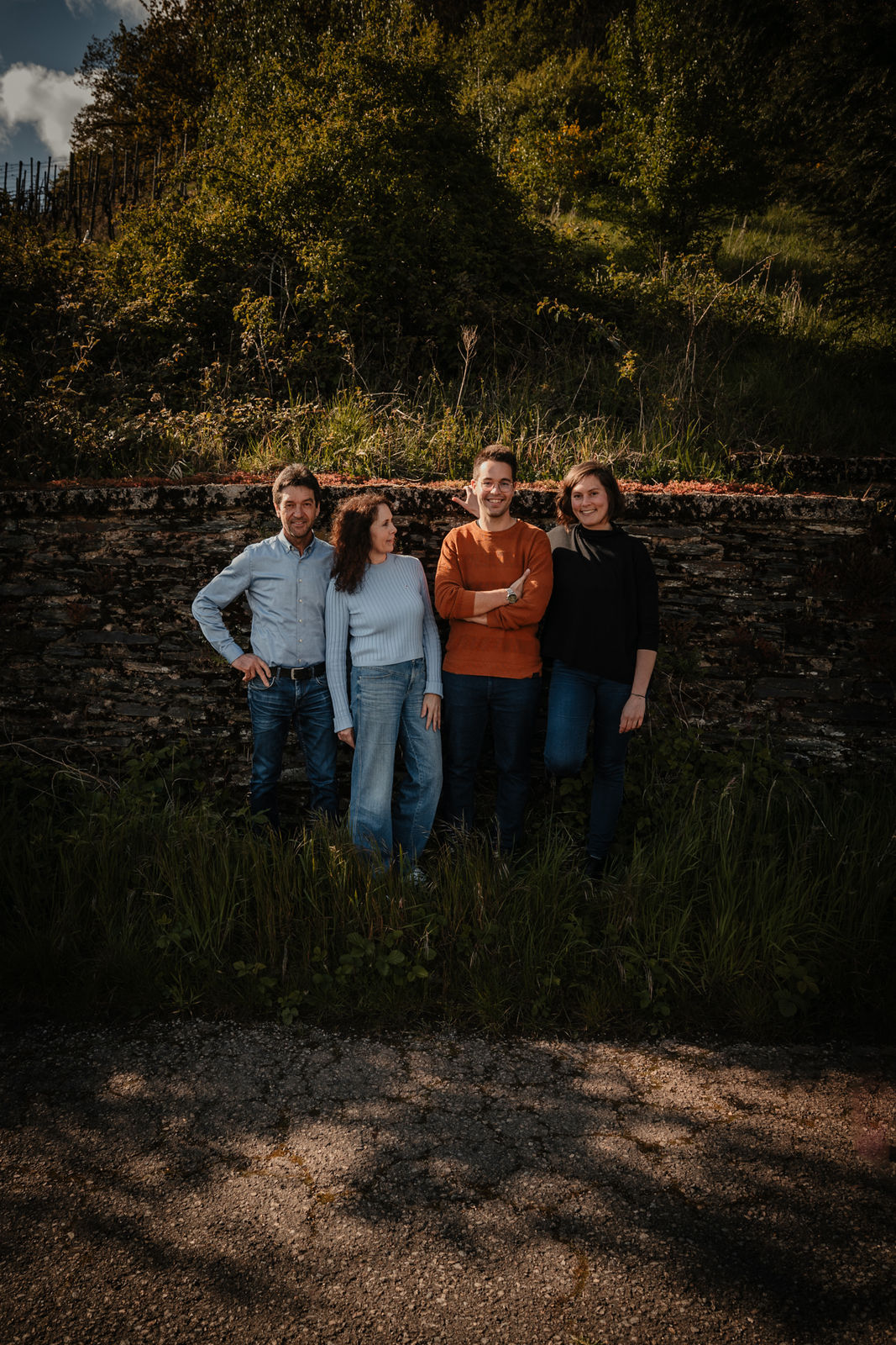 Vier Personen (Familie Gindorf) stehen vor einer alten Schiefersteinmauer in den Weinbergen von Schweich; Gruppenfoto im warmen Abendlicht, umgeben von grüner Natur – Weingut Günter Gindorf.