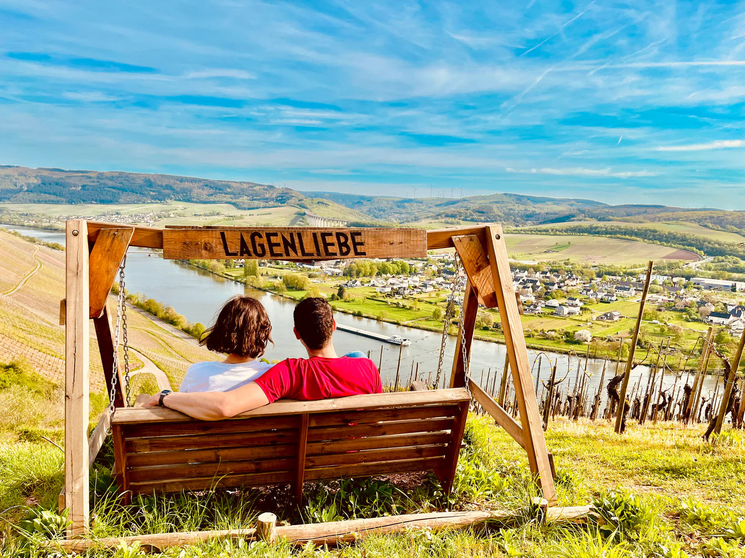 Zwei Personen sitzen auf der Lagenliebe-Schaukel in den Mosel-Steillagen oberhalb von Schweich und genießen den weiten Blick über Weinberge, Fluss und Tal bei sonnigem Wetter. (Annaberg Lagenliebe Aussichtspunkt)