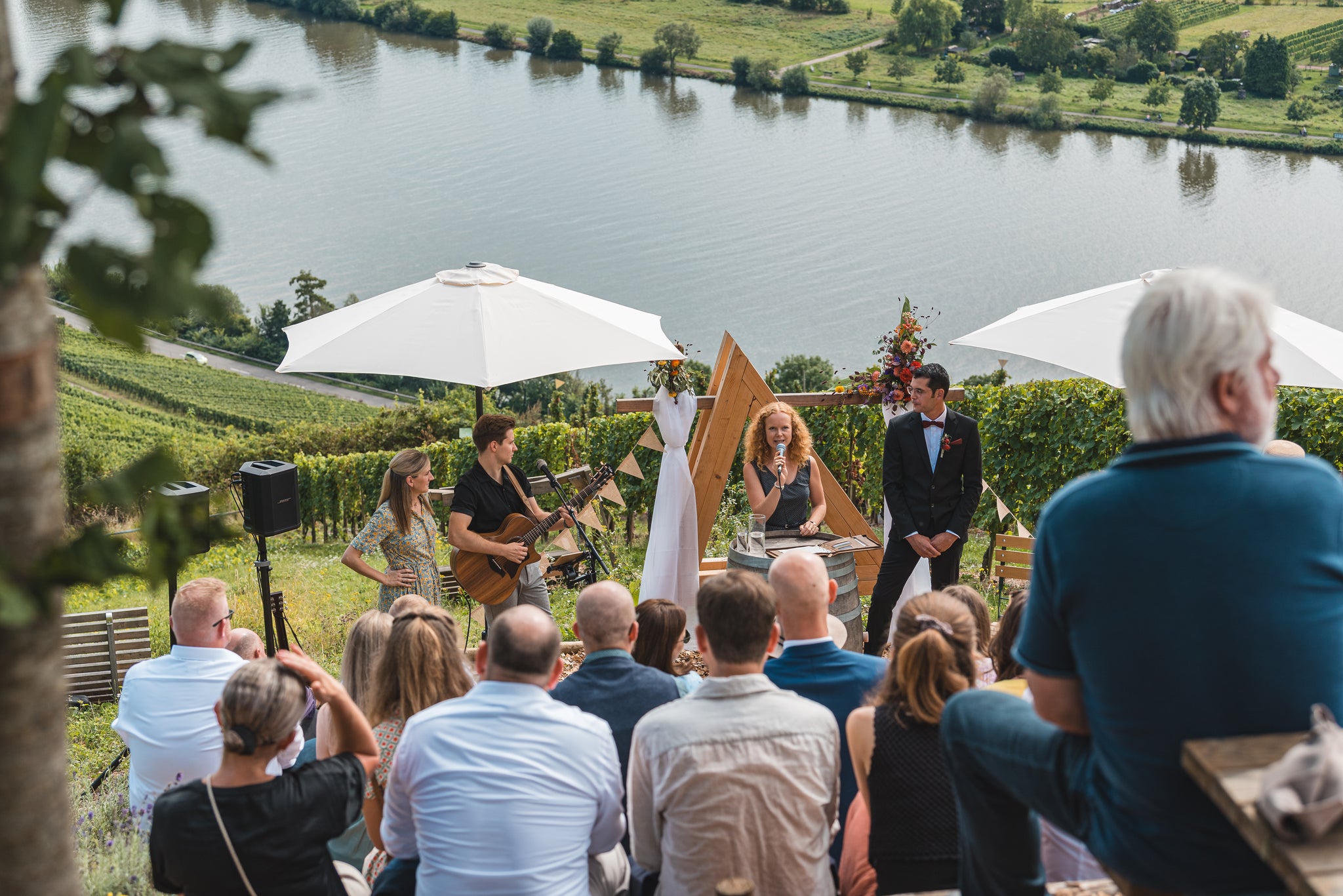 Freie Trauung in den Weinbergen oberhalb der Mosel – Gäste sitzen auf terrassenartigen Stufen, vor ihnen ein dekorierter Traubogen, Musiker und Rednerin, mit Panoramablick über das Moseltal