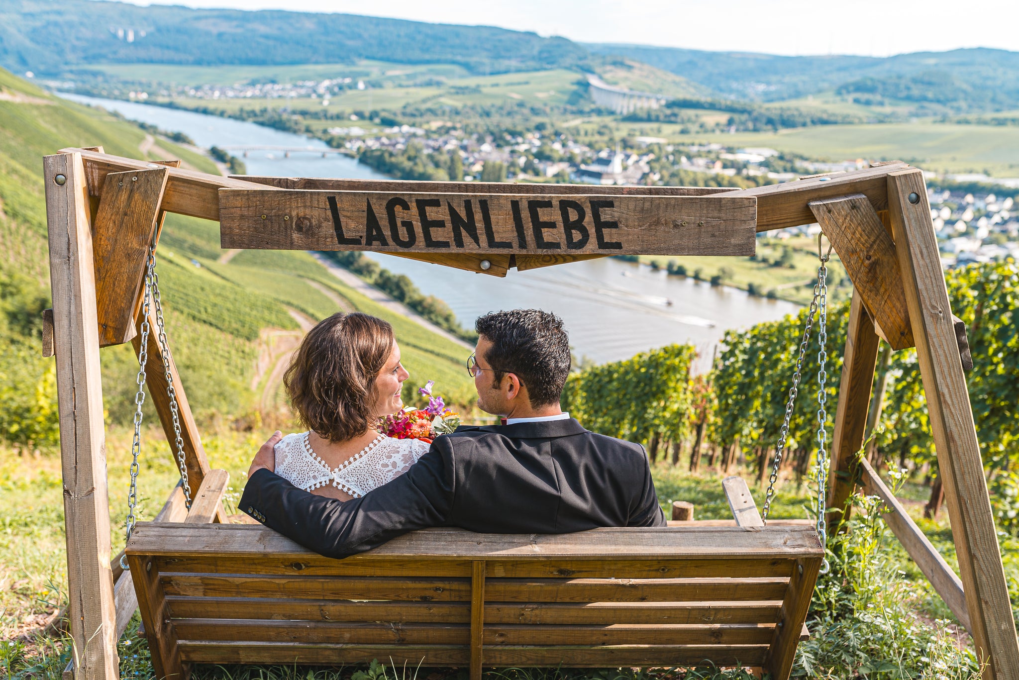 Paar auf Holzschaukel mit der Aufschrift ‚Lagenliebe‘ in den Weinbergen oberhalb von Schweich – romantischer Blick über Mosel, Rebhänge und das Moseltal.